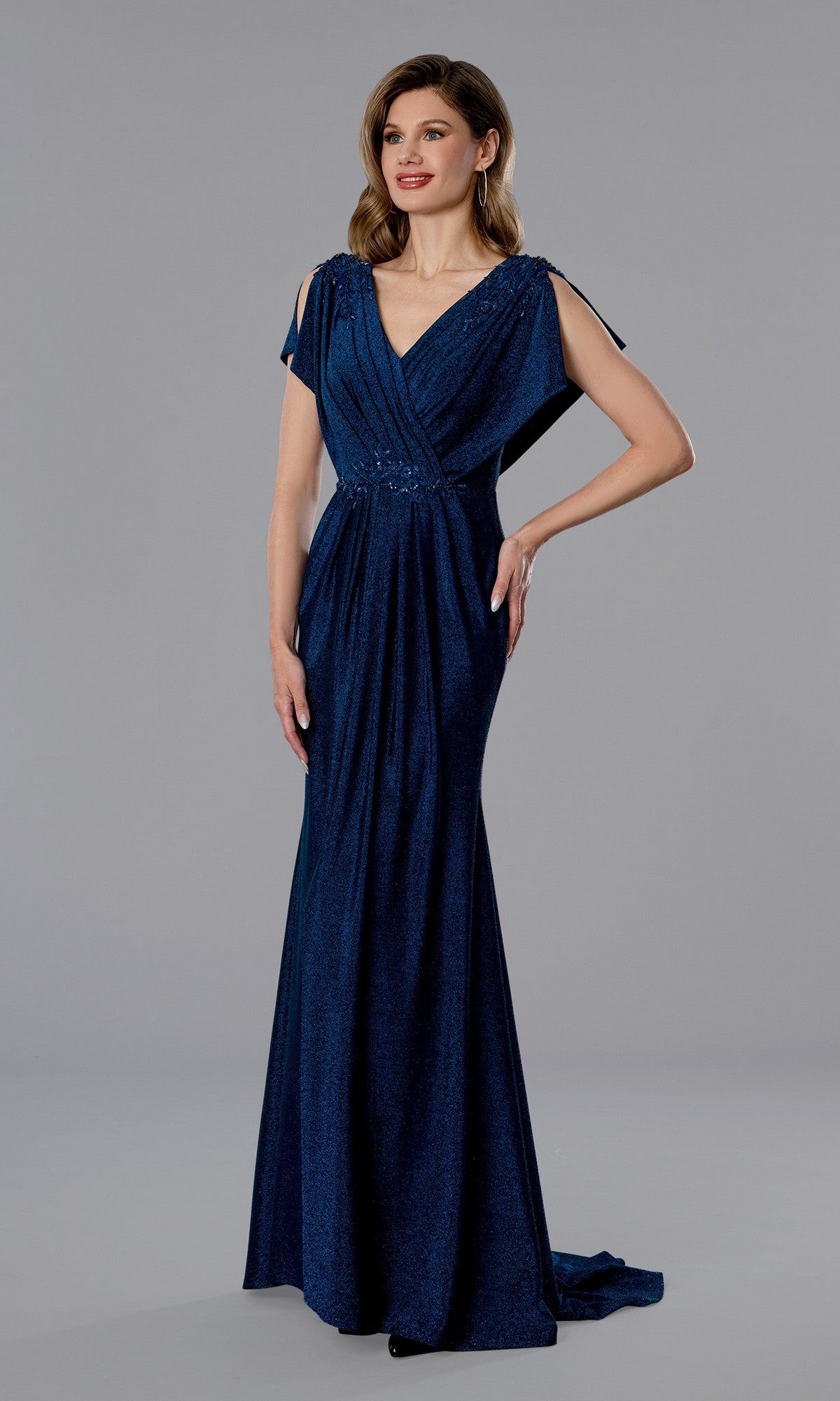 A woman in a blue Stella Couture 25350 long formal dress with a v-neck and short draped sleeves stands against a plain gray background.